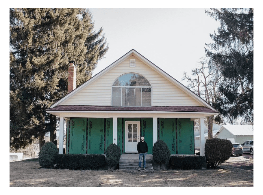 A man stands in front of a two-story house with a pitched roof, showcasing the work of a Spokane general contractor. The house features large green panels on its facade, flanked by two evergreen trees and several bushes lining the front. The scene is bathed in daylight.
