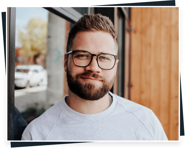 A man with short hair and a beard, who could be Joshua Johnson, is wearing glasses and a light gray sweatshirt. He stands outside, leaning against a glass window. The background features a blurry view of cars and a wooden wall.