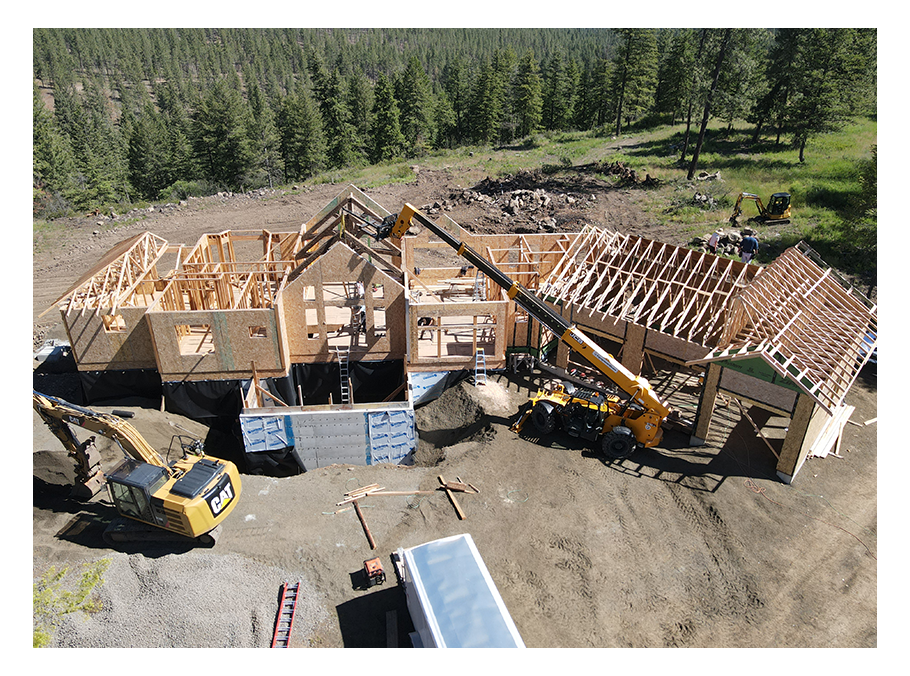 Aerial view of a new residential construction featuring a wooden house in a forested area. Cranes and machinery surround the site, with building materials scattered nearby. The structure's frame is partially complete, showcasing progress in the build.