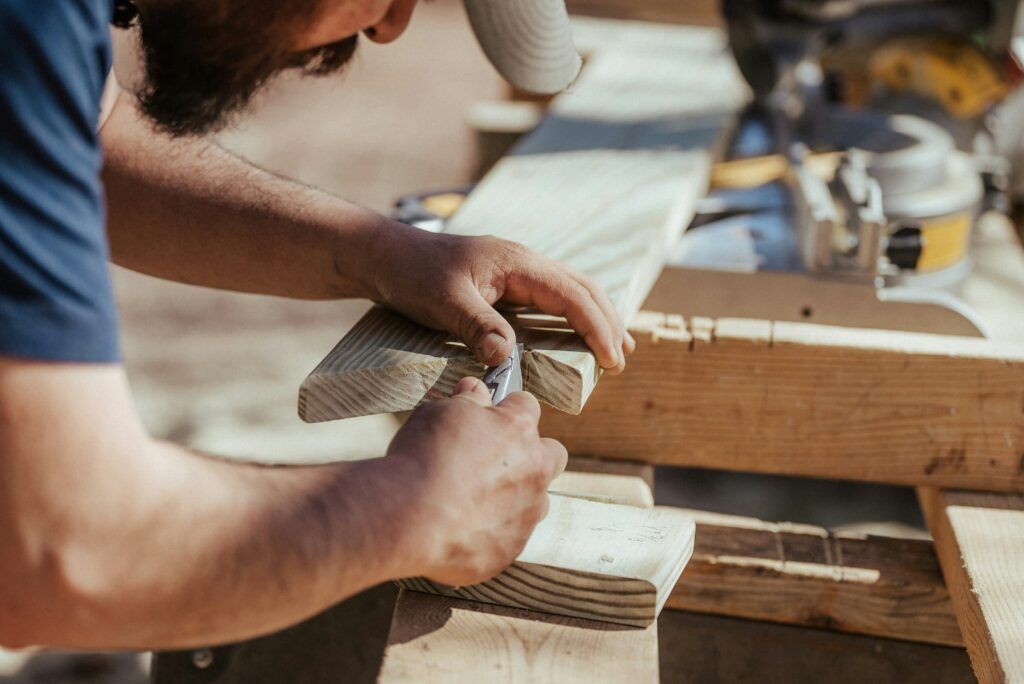 A person wearing a cap is carefully carving a piece of wood with a knife. A contractor's saw rests in the background on a wooden workbench. The outdoor setting emphasizes the skilled woodworking activity taking place.