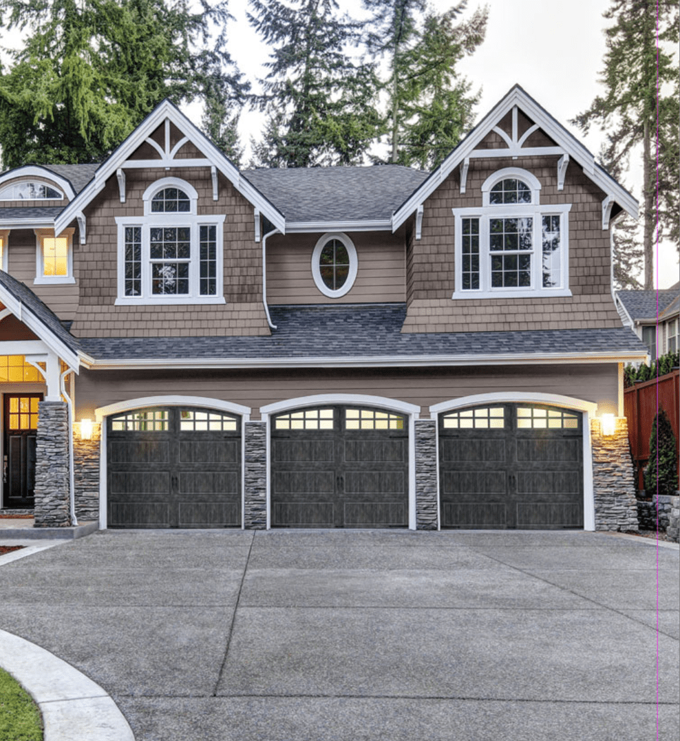 A two-story suburban house with gray stone and wood siding, featuring four dark garage doors, large windows, exterior lights, and surrounded by trees and a concrete driveway.