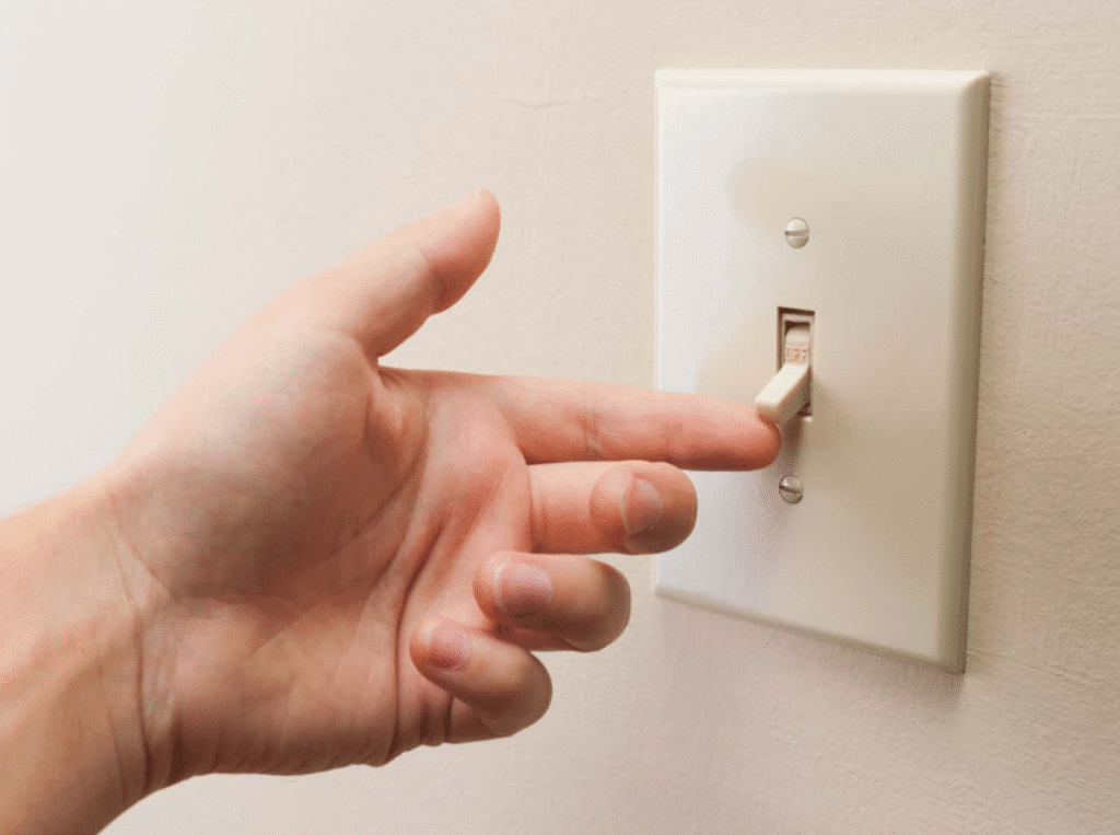 A left hand reaches toward a light switch on a white wall, with the index finger extended and about to flip the beige switch to the off position.
