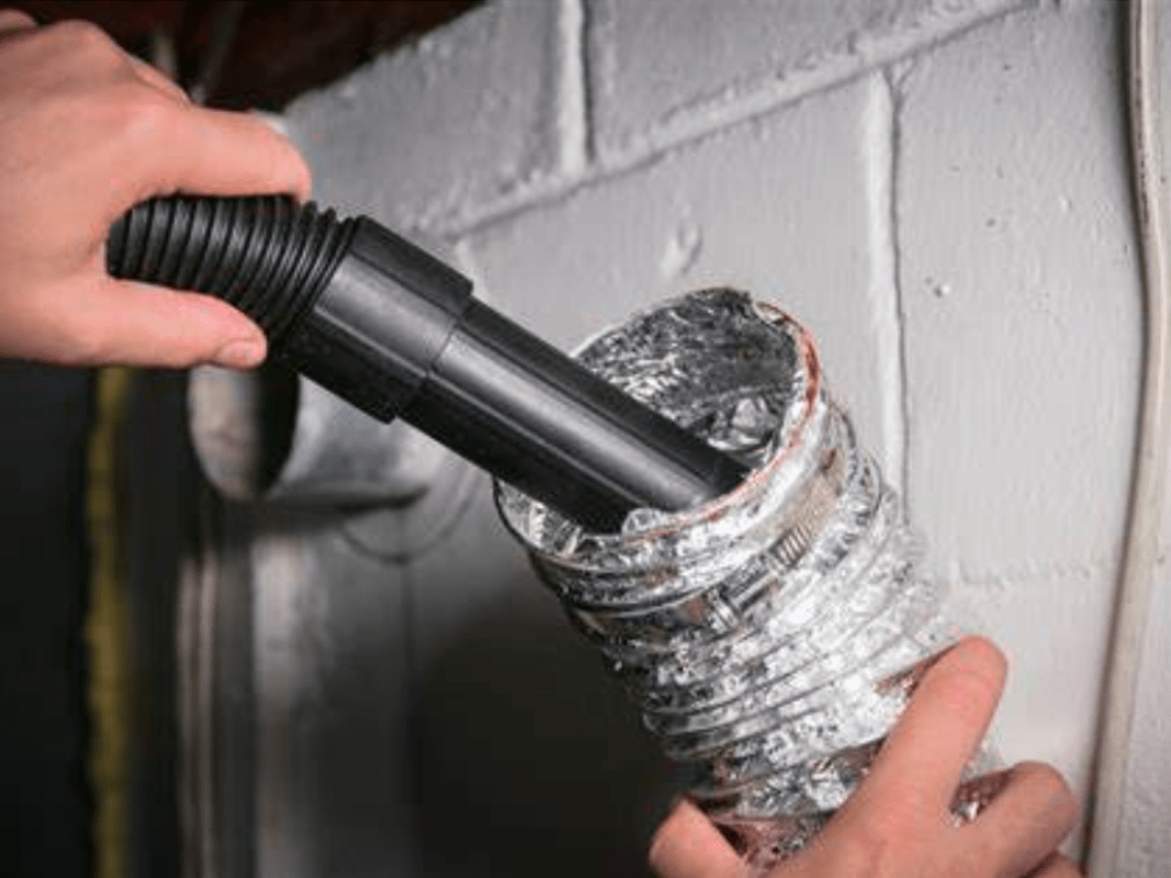 A person holds a flexible dryer vent duct while using a vacuum cleaner hose to clean inside the duct near a white painted brick wall.